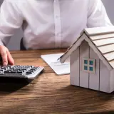 Persona calculando el valor de una propiedad, con una maqueta de casa, calculadora y documentos. Aprende como tasar una vivienda.