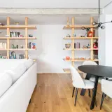 Modern living room with white sofa, wooden shelves filled with books and decor, and a dining area. Un ejemplo de cambio de uso de local a vivienda.