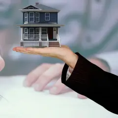 Mano sostiene casa miniatura, mientras otra firma documentos. Posiblemente se trate de gastos de cancelación de hipoteca.