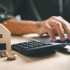 Calculadora, monedas y casa de madera junto a un portátil. Símbolos de porcentaje flotan, sugiriendo el cálculo del precio vivienda.