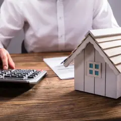 Persona calculando el valor de una propiedad, con una maqueta de casa, calculadora y documentos. Aprende como tasar una vivienda.