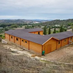 Edificio de madera en forma de L, que es una finca rústica, rodeado de vegetación y colinas.