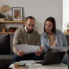 Pareja revisa documentos y laptop, debatiendo qué tasadora tasa más alto para su hipoteca.