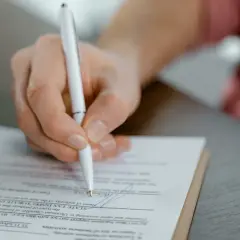 Mano firmando un documento, posiblemente un acuerdo de dación en pago entre particulares, con un bolígrafo blanco sobre papel.