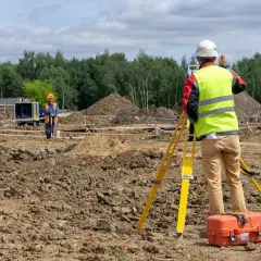 Topógrafo con equipo de levantamiento topográfico en obra. Otro trabajador supervisa el terreno.