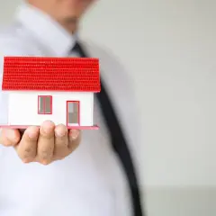 Un hombre con corbata negra sostiene una maqueta de casa con techo rojo. Representa la ley de arrendamientos urbanos.