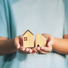 Manos sostienen dos casas de madera, como si fueran un expediente de dominio.