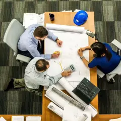 Tres personas analizan planos en una mesa. El significado de este predio se discute con una calculadora y un casco azul.