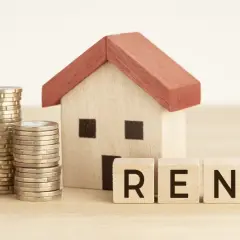 Casa de juguete con monedas y cubos que dicen "RENT". Representa el acuerdo entre arrendador y arrendatario.