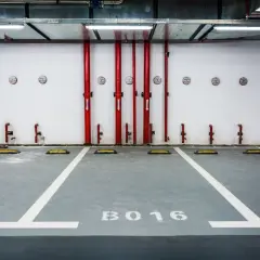 Plaza de garaje B016, con líneas blancas y topes amarillos. Tuberías rojas y luces en el techo. Ideal para tasar plaza de garaje.