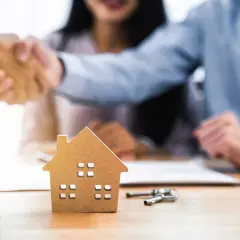 Pareja estrecha manos con agente inmobiliario. Una maqueta de casa y llaves sobre la mesa sugieren que las arras se descuentan de la hipoteca.