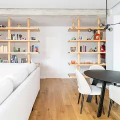 Modern living room with white sofa, wooden shelves filled with books and decor, and a dining area. Un ejemplo de cambio de uso de local a vivienda.