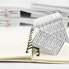 Casa de papel con números, simulando un código registral único, sobre un cuaderno abierto y pilas de documentos.