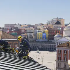 Dos operarios con arnés y casco amarillo trabajan en un tejado, con la ciudad de Nápoles de fondo. Tienen el permiso para cambiar tejado.
