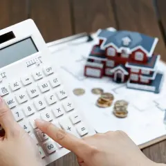 Manos calculando con calculadora blanca, casa modelo, monedas y bolígrafo. Planificando cómo desgravar alquiler.