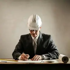 Un aparejador que es, con casco y traje, dibuja planos en su escritorio.