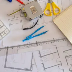 Herramientas de diseño sobre una superficie gráfica: teclado, cinta métrica, tijeras, compás, regla, cuaderno y lápiz.