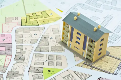 Maqueta de vivienda sobre planos urbanísticos. El modelo representa un edificio de varias plantas, con balcones y ventanas. Los planos detallan parcelas y edificaciones, sugiriendo la planificación para determinar el **valor catastral vivienda**.