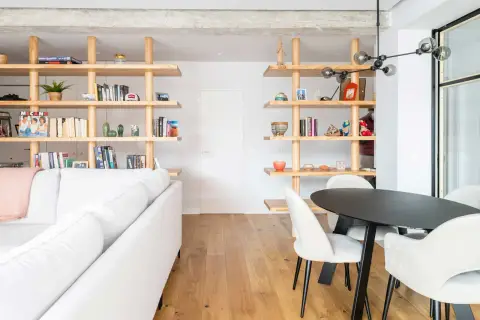 Modern living room with white sofa, wooden shelves filled with books and decor, and a dining area. Un ejemplo de cambio de uso de local a vivienda.