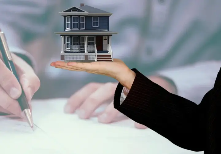 Mano sostiene casa miniatura, mientras otra firma documentos. Posiblemente se trate de gastos de cancelación de hipoteca.