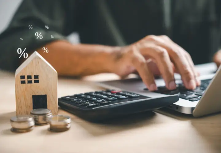 Calculadora, monedas y casa de madera junto a un portátil. Símbolos de porcentaje flotan, sugiriendo el cálculo del precio vivienda.