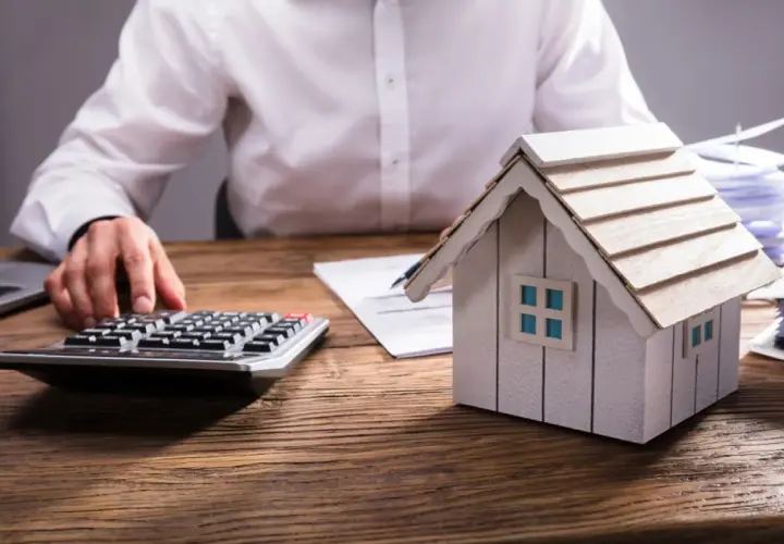 Persona calculando el valor de una propiedad, con una maqueta de casa, calculadora y documentos. Aprende como tasar una vivienda.