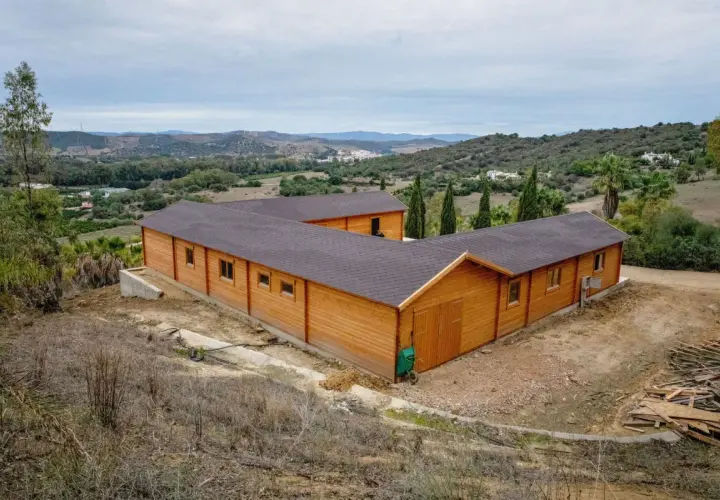 Edificio de madera en forma de L, que es una finca rústica, rodeado de vegetación y colinas.