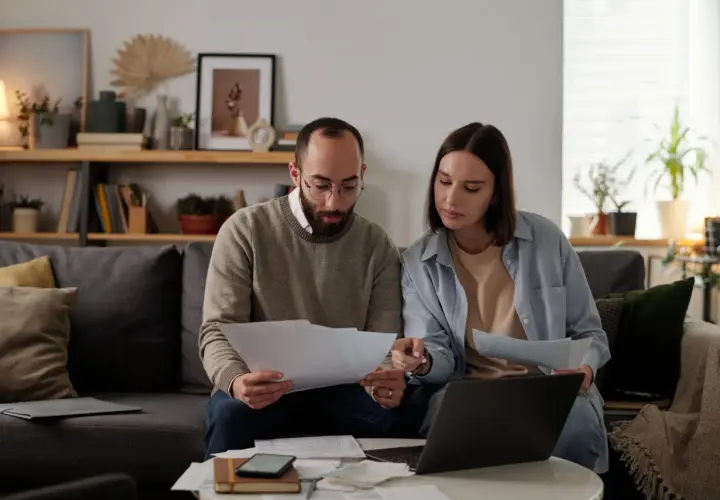 Pareja revisa documentos y laptop, debatiendo qué tasadora tasa más alto para su hipoteca.