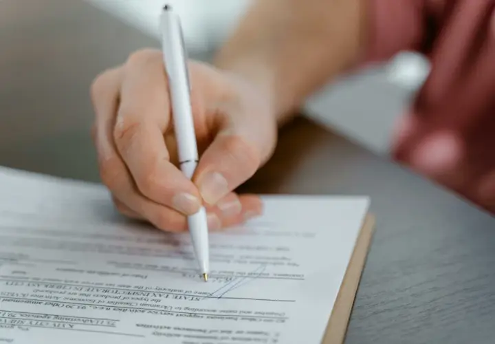 Mano firmando un documento, posiblemente un acuerdo de dación en pago entre particulares, con un bolígrafo blanco sobre papel.