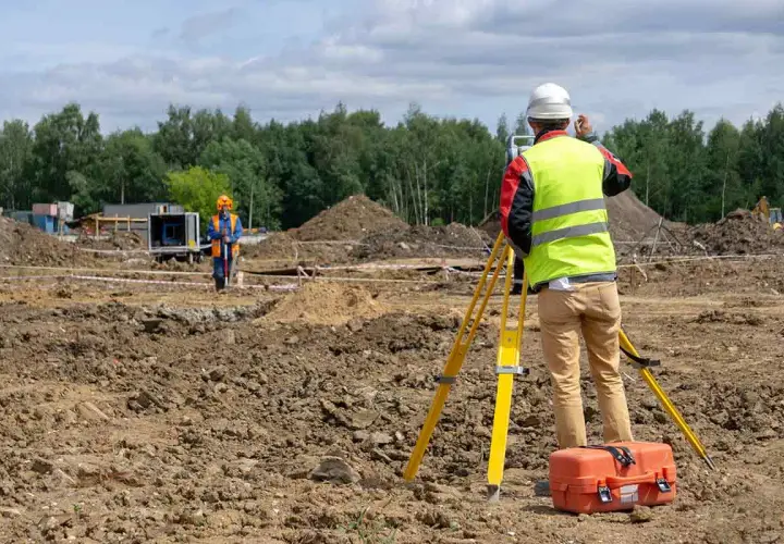 Topógrafo con equipo de levantamiento topográfico en obra. Otro trabajador supervisa el terreno.