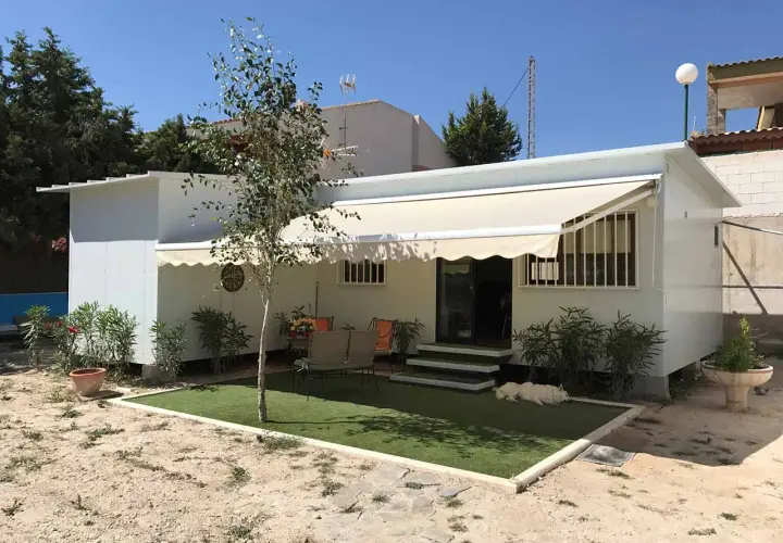 Una moderna casa de aperos blanca con toldo, césped artificial y un perro blanco descansando.