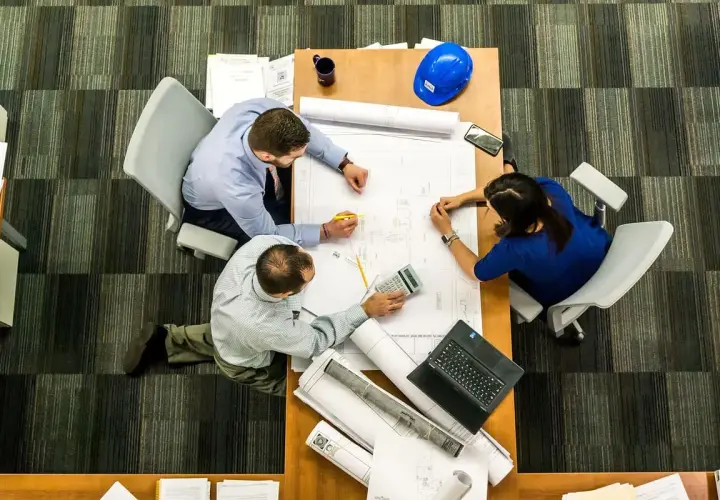 Tres personas analizan planos en una mesa. El significado de este predio se discute con una calculadora y un casco azul.