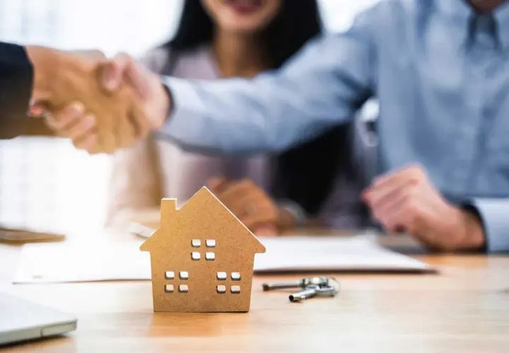 Pareja estrecha manos con agente inmobiliario. Una maqueta de casa y llaves sobre la mesa sugieren que las arras se descuentan de la hipoteca.