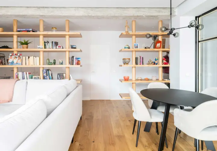 Modern living room with white sofa, wooden shelves filled with books and decor, and a dining area. Un ejemplo de cambio de uso de local a vivienda.