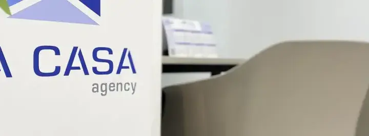 Logo de "LA CASA agency" en un cartel blanco, con sillas de oficina al fondo. Buscando la casa agency opiniones.