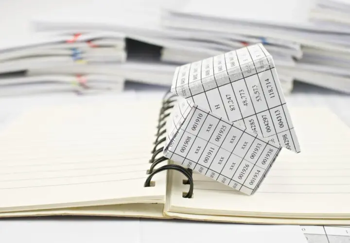 Casa de papel con números, simulando un código registral único, sobre un cuaderno abierto y pilas de documentos.