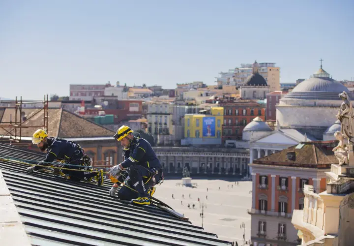 Dos operarios con arnés y casco amarillo trabajan en un tejado, con la ciudad de Nápoles de fondo. Tienen el permiso para cambiar tejado.