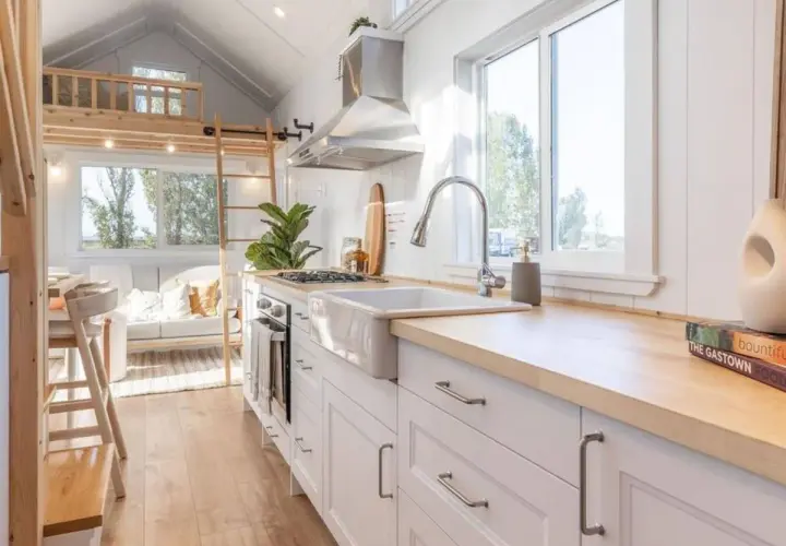 Cocina luminosa y funcional de una tiny house, con encimeras de madera, fregadero blanco y una acogedora sala de estar al fondo.
