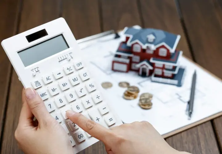 Manos calculando con calculadora blanca, casa modelo, monedas y bolígrafo. Planificando cómo desgravar alquiler.