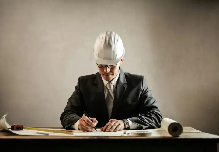 Un aparejador que es, con casco y traje, dibuja planos en su escritorio.