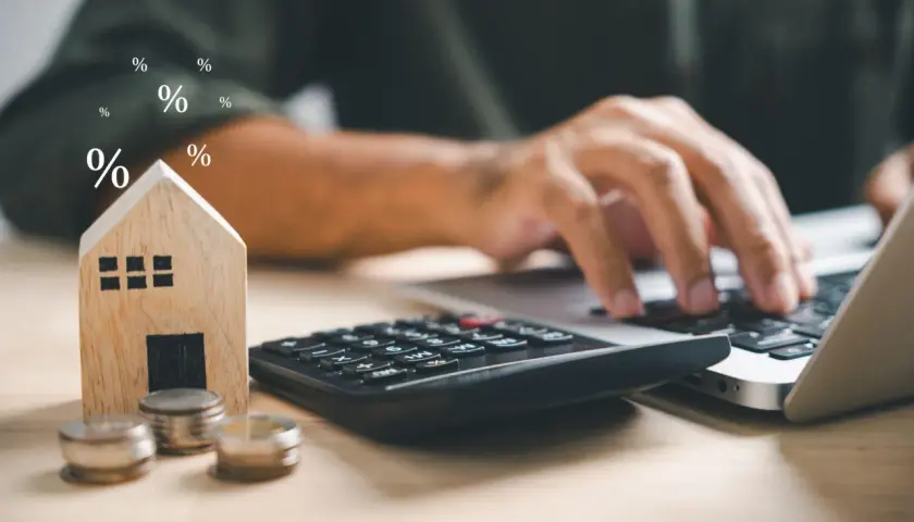 Calculadora, monedas y casa de madera junto a un portátil. Símbolos de porcentaje flotan, sugiriendo el cálculo del precio vivienda.