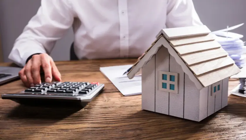 Persona calculando el valor de una propiedad, con una maqueta de casa, calculadora y documentos. Aprende como tasar una vivienda.