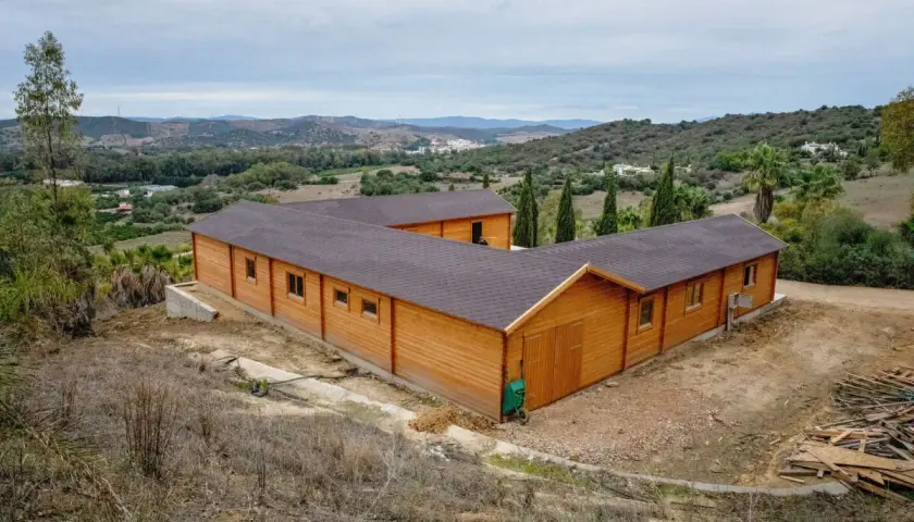 Edificio de madera en forma de L, que es una finca rústica, rodeado de vegetación y colinas.