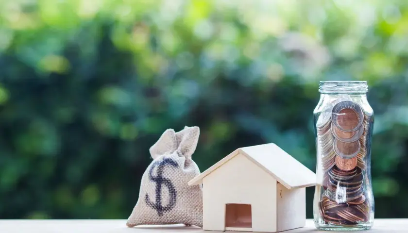 Saco de dinero, casa de madera y frasco lleno de monedas, simbolizando el pleno dominio de tus finanzas para adquirir tu hogar.