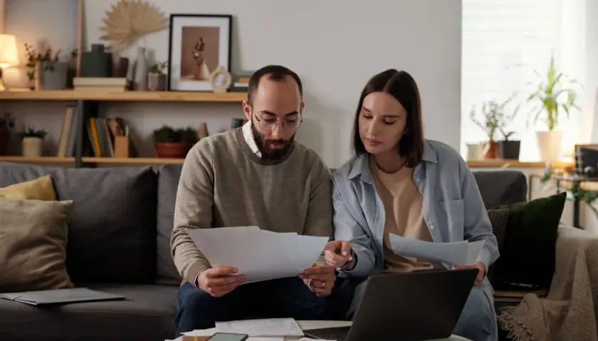Pareja revisa documentos y laptop, debatiendo qué tasadora tasa más alto para su hipoteca.