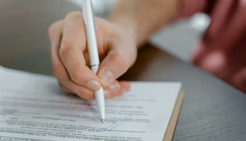 Mano firmando un documento, posiblemente un acuerdo de dación en pago entre particulares, con un bolígrafo blanco sobre papel.