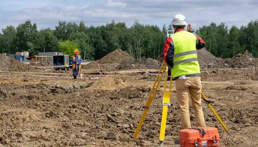 Topógrafo con equipo de levantamiento topográfico en obra. Otro trabajador supervisa el terreno.