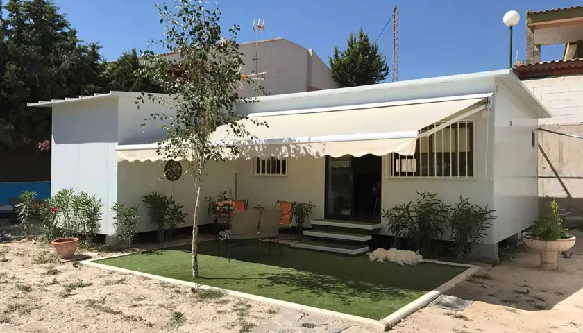 Una moderna casa de aperos blanca con toldo, césped artificial y un perro blanco descansando.