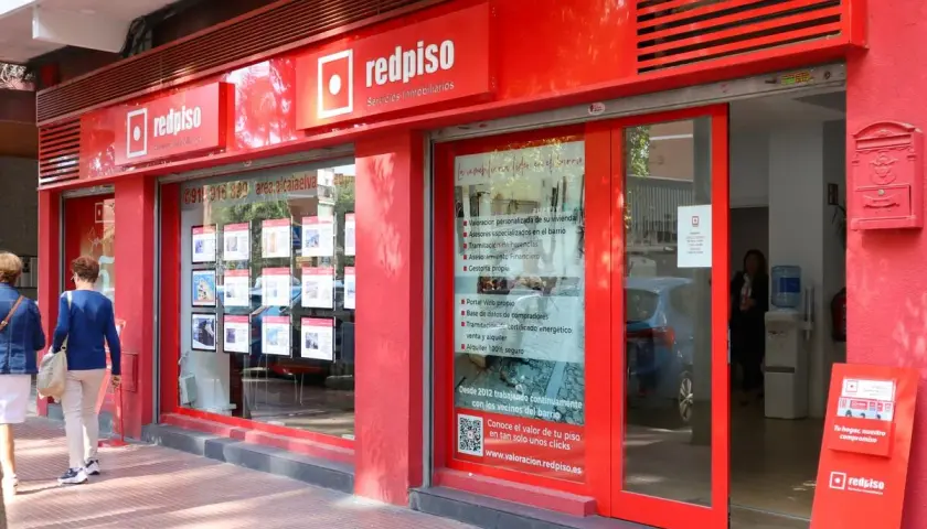 Redpiso, inmobiliaria con opiniones positivas. Fachada roja, escaparate con anuncios de pisos y dos mujeres mirando.
