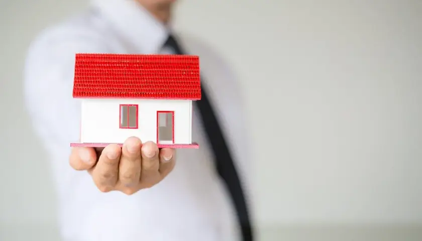 Un hombre con corbata negra sostiene una maqueta de casa con techo rojo. Representa la ley de arrendamientos urbanos.